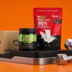 Embalagens de Whey Protein 80% e Creatina da Growth Supplements sobre suporte com caixa de entrega Amazon Prime e halteres em estúdio laranja