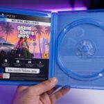 Mão segurando um estojo azul de PlayStation 5 aberto com a bandeja de disco totalmente vazia e um encarte de GTA 6 com o aviso NO DISC INCLUDED e voucher de download